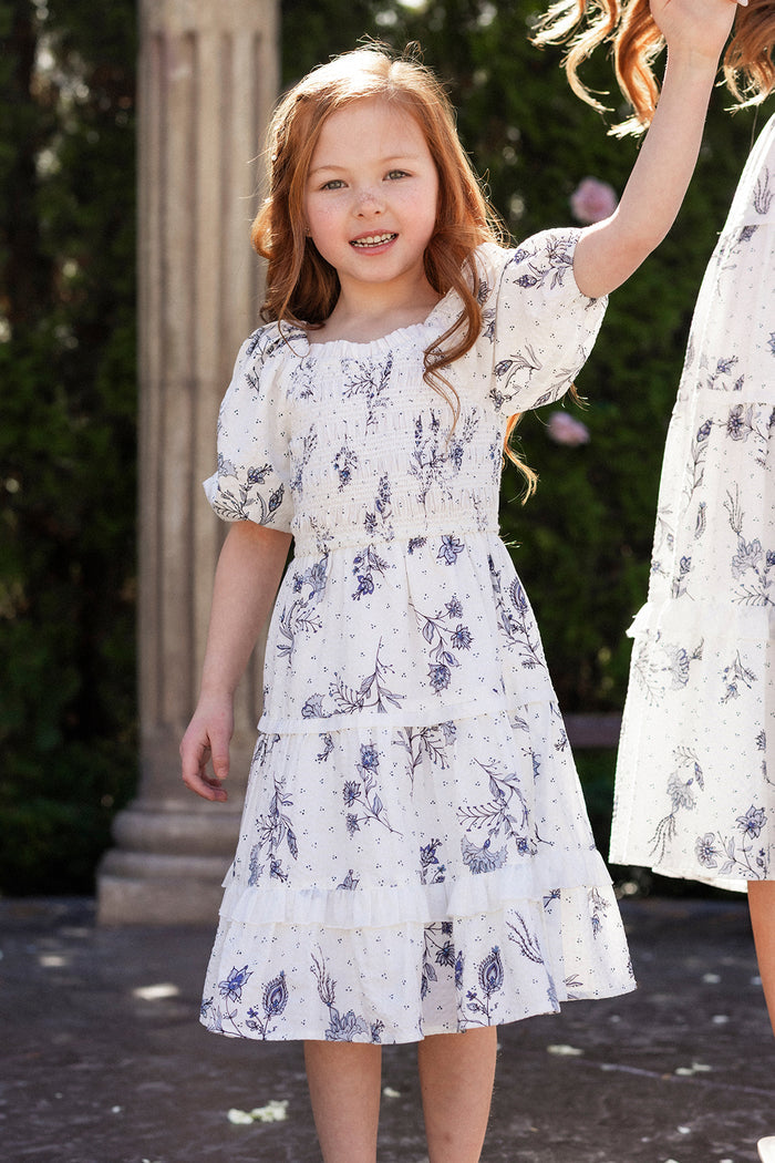 A young girl with long red hair smiles outdoors in the Ivy City Co Mini Roselyn Dress in Blue Enchanted Floral, standing near another person in a similar spring dress with greenery and a stone column behind them.