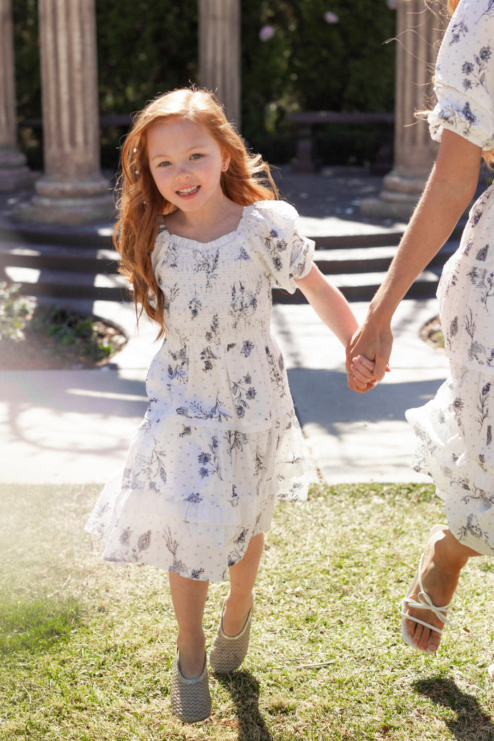 A young girl with long red hair smiles while holding an adult’s hand, both wearing matching Mini Roselyn Dresses in Blue Enchanted Floral by Ivy City Co, walking outside on grass in a garden-like setting.