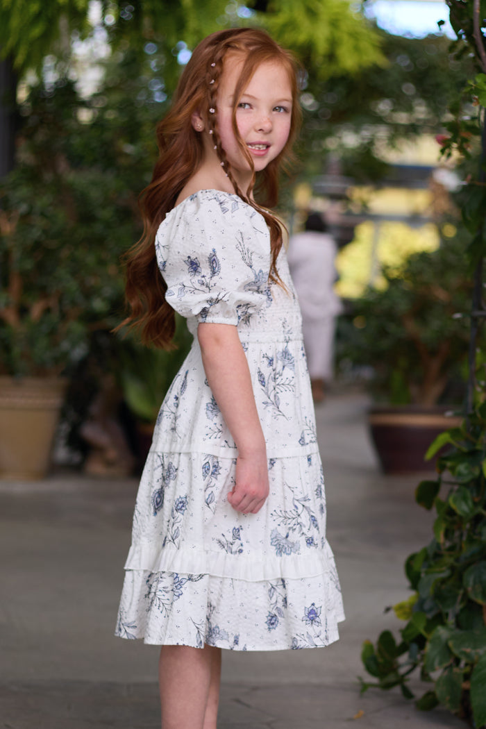 A young girl with long red hair stands sideways, smiling slightly and surrounded by greenery, wearing the Ivy City Co Mini Roselyn Dress in Blue Enchanted Floral in a lush garden or greenhouse setting.
