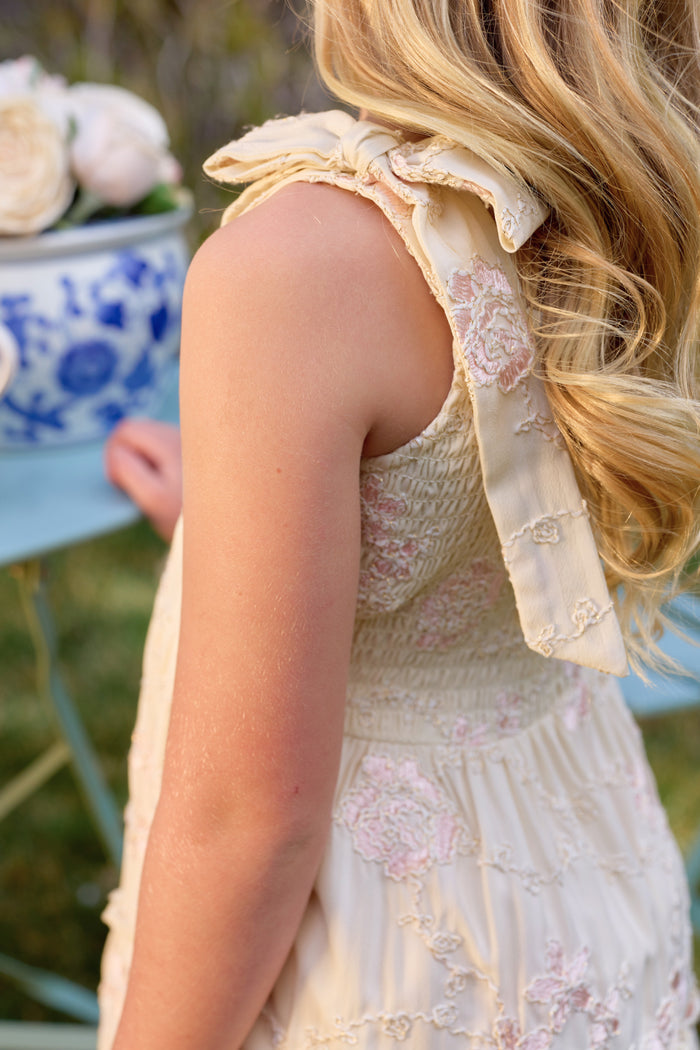 A young girl with long, wavy blonde hair wears the Ivy City Co Mini Tara Dress in Gilded Rose Stripe with a shoulder bow, standing by a blue table holding a floral arrangement in a white-and-blue vase.