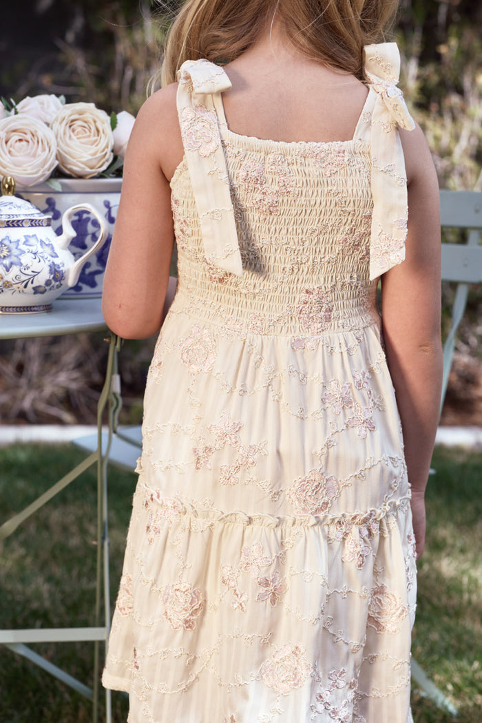 A young girl wearing the Ivy City Co Mini Tara Dress in Gilded Rose Stripe, adorned with bows on the straps, stands outdoors near a table set with white roses and a blue teapot.