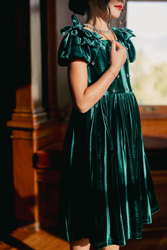 A young girl wears the Ivy City Co Mini Tiara Dress in Green Velvet with puffed sleeves, standing indoors by a window. She holds her necklace with both hands and her face, partially visible, is highlighted by red lipstick.