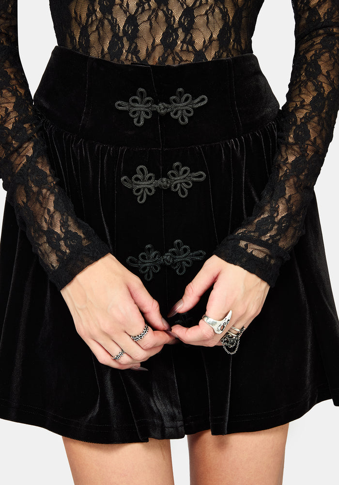 A model wearing a black gothic-style flared velvet mini skirt with bat-wing shaped front buttons, paired with black platform boots, against a light gray background.