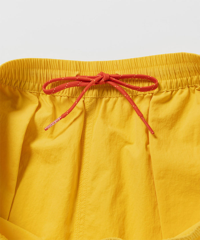 Close-up of banana colored Gramicci Nylon Reef Shorts with a red drawstring on a light-tone background.
