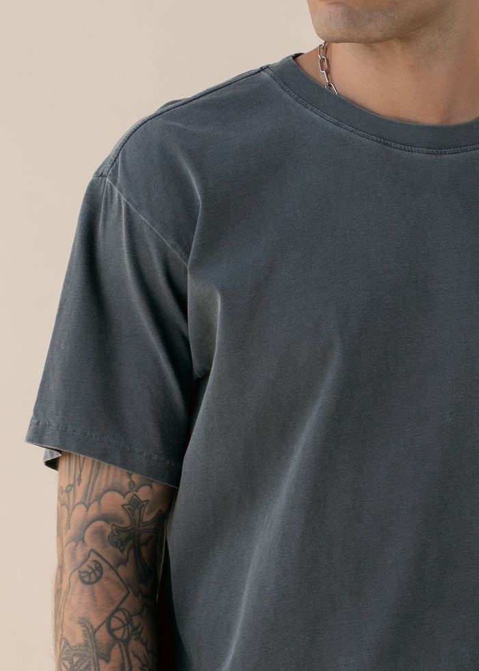 Close up of person wearing a dark gray t-shirt with a tattooed arm on a beige background