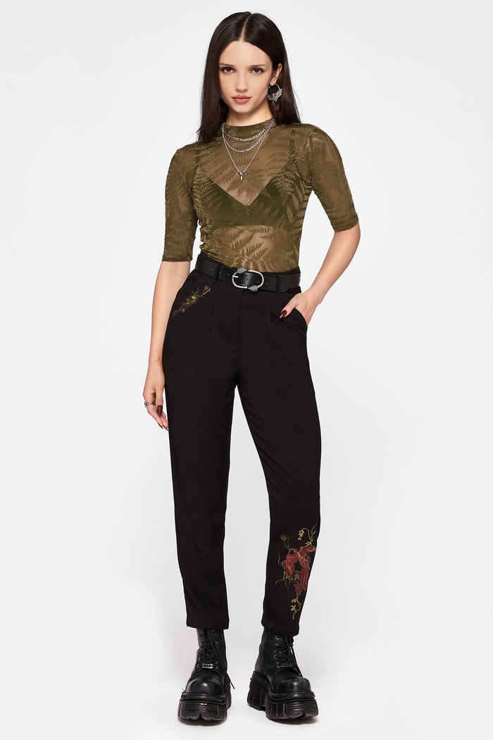 Woman wearing a green top and black pants with floral embroidery on a white background