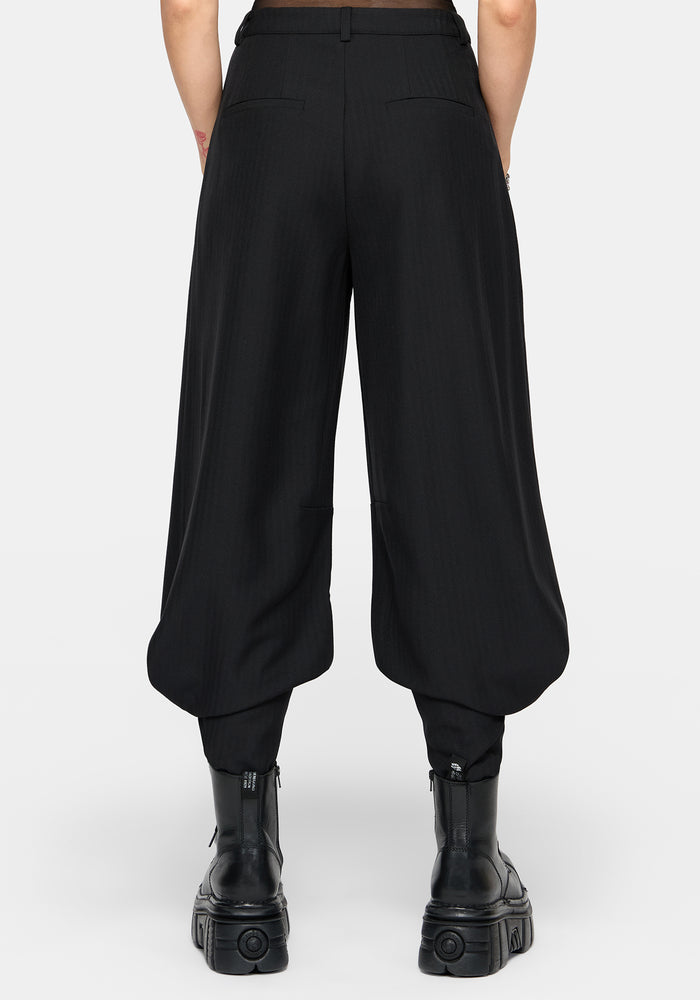 Rear view of a model wearing black wide-leg balloon pants with gathered legs and black platform ankle boots against a light gray studio background.