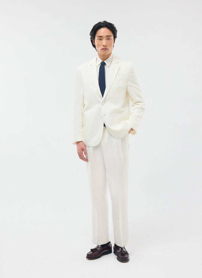 A model wears the full Percival Relaxed Tailored Linen Suit in white, styled with a navy tie and brown loafers.