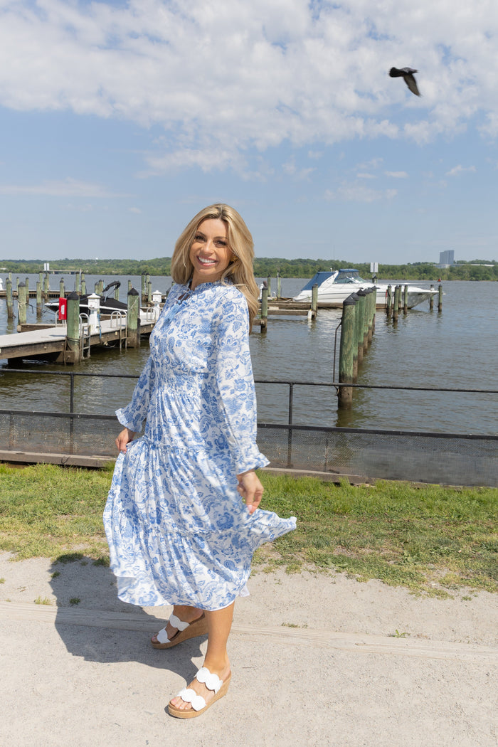 Woman in a blue dress standing by a waterfront with boats and a clear sky.