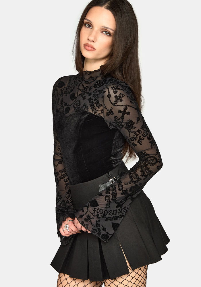 A woman modeling a black long-sleeve top with sheer floral lace sleeves and a pleated mini skirt, paired with fishnet tights against a light gray studio background.