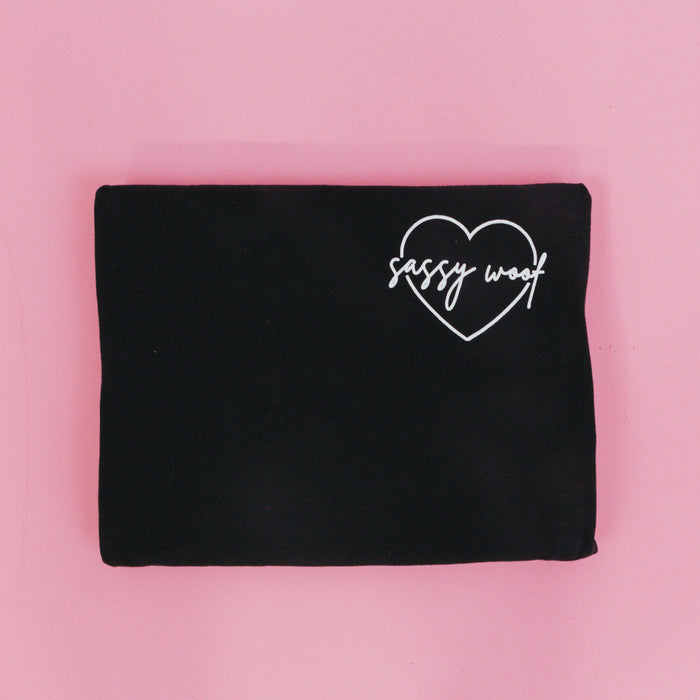 The Sassy Woof Branded Tee by Sassy Woof, a folded black lightweight shirt with a white heart outline and "sassy woof" inside, is displayed on a pink background.