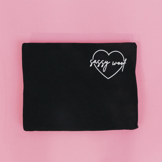 The Sassy Woof Branded Tee by Sassy Woof, a folded black lightweight shirt with a white heart outline and "sassy woof" inside, is displayed on a pink background.