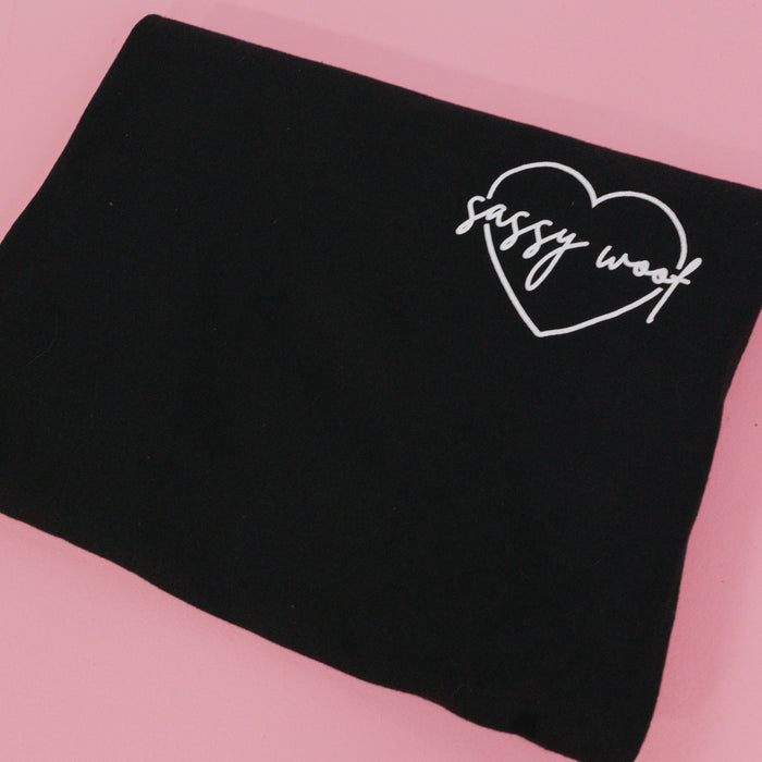 The Sassy Woof Branded Tee by Sassy Woof is a folded black 100% cotton t-shirt featuring a white heart outline with "sassy woof" inside, displayed on a pink background.