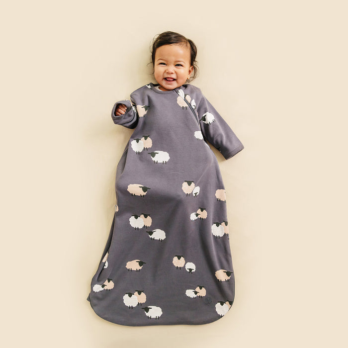 Baby wearing a gray/indigo sleep bag with sleeves in sheep pattern on a beige background