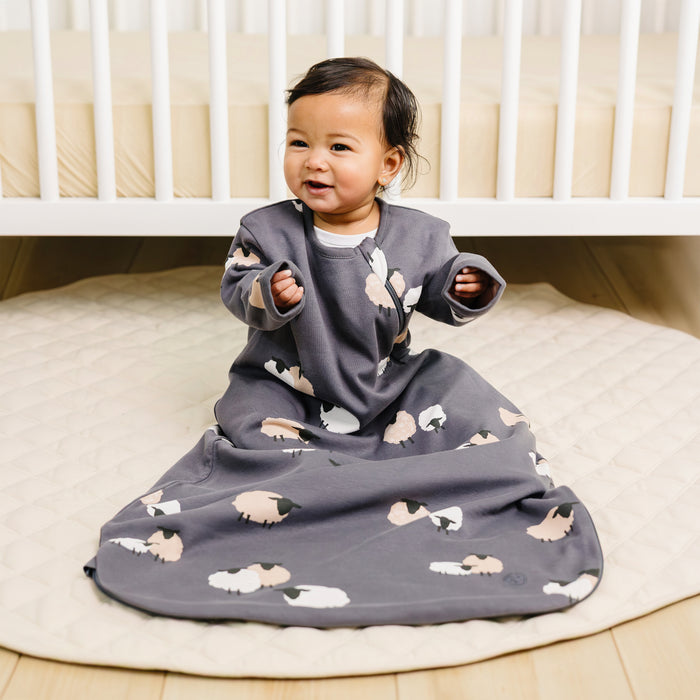 Baby in a gray /indigo sleep sack with sleeves with sheep prints, sitting on a baby rug.