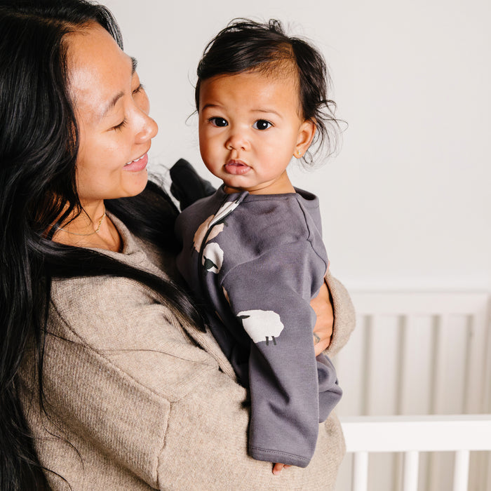 A woman holding a baby wearing a gray sleep sack with sheep design indoors.
