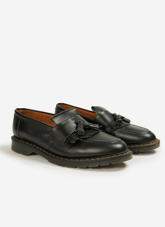A pair of Percival x Solovair black hi-shine leather tassel loafers with a chunky rubber sole on a white background.