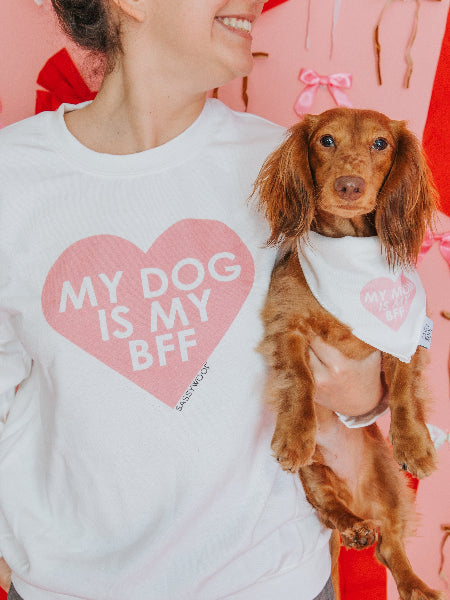 A woman in a Sassy Woof "My Dog is my BFF" pink sweatshirt holds her small brown dog in a matching bandana, both smiling against a pink backdrop with red and pink ribbons, showcasing their special bond.
