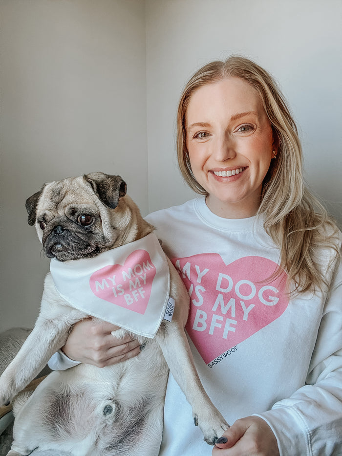 A smiling woman wears a pink "My Dog is my BFF" sweatshirt by Sassy Woof, sitting beside a pug in a matching bandana that says "My Mom is my BFF." Both pose happily in a brightly lit room.