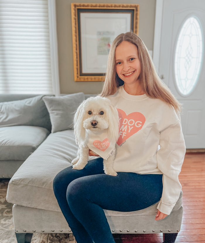 A smiling woman sits on a couch with her small white dog. Both wear Sassy Woof “My Dog is my BFF” (PINK) sweatshirts, showing off their matching style and love for each other.