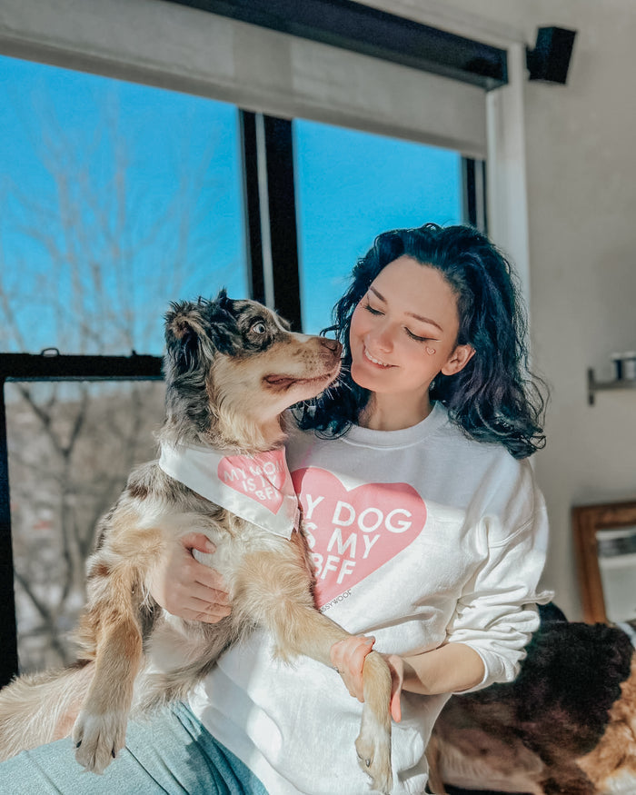 A woman smiles, holding her Australian Shepherd. She wears the Sassy Woof “My Dog is my BFF” pink sweatshirt, while her pup sports a matching bandana. Sunlight pours through a large window behind them.