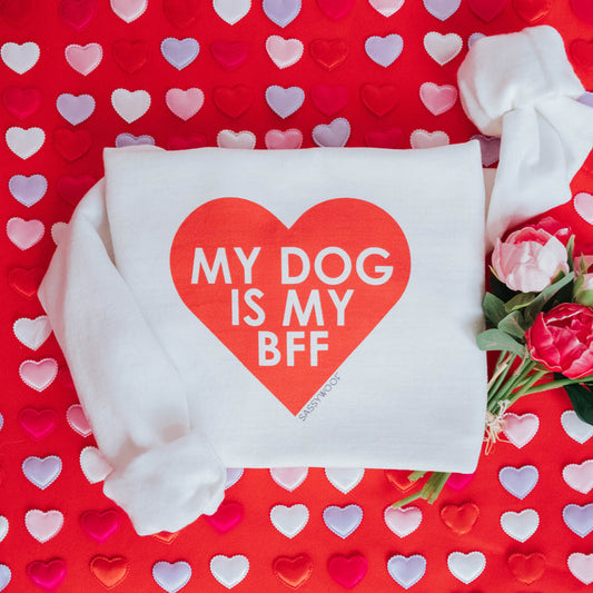 The Sassy Woof "Sweatshirt - My Dog is my BFF (RED)" features a bold red heart and fun text on white, displayed on a red backdrop with colorful hearts and tulips beside this cozy unisex crewneck.