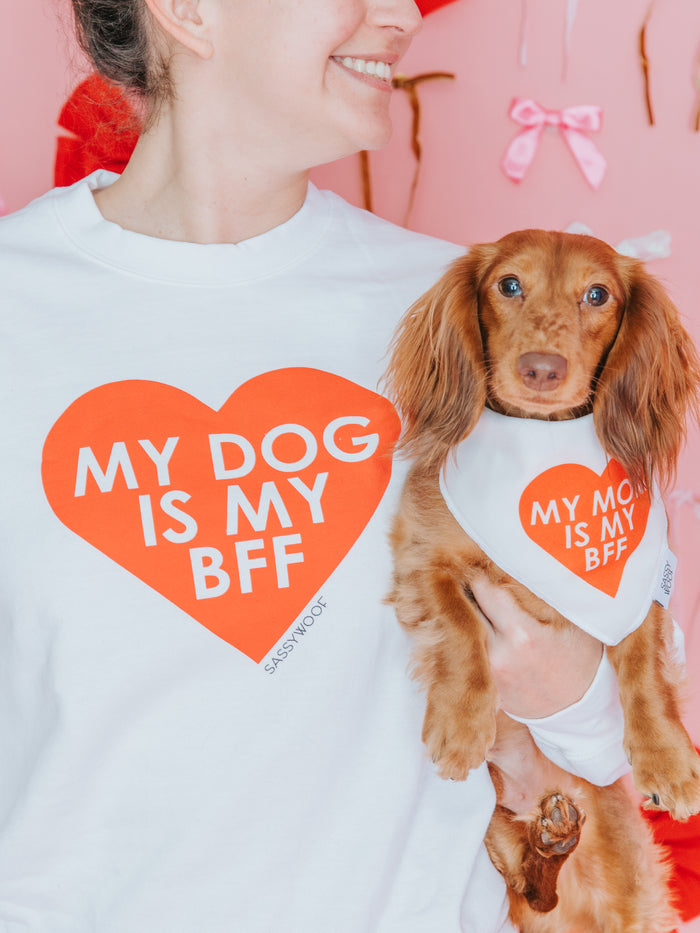 A person wears the Sassy Woof Sweatshirt - My Dog is my BFF (RED) and stands next to a brown dog in a matching "My Mom is My BFF" bandana, with pink bows and flowers blurred in the background.