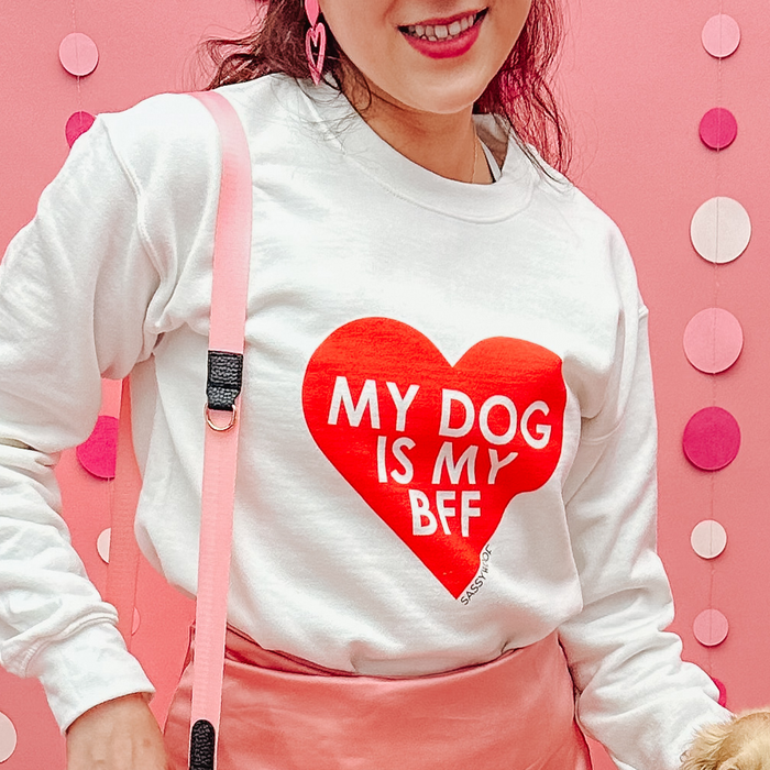 A woman smiles in front of a pink background, wearing the Sassy Woof Sweatshirt - My Dog is my BFF (RED), a unisex white crewneck featuring a large red heart and bold lettering. She pairs it with pink earrings and a strap.
