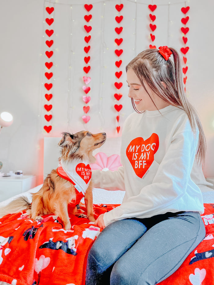 A woman wears the Sassy Woof "My Dog is my BFF (RED)" sweatshirt, sitting with her dog in a matching bandana. Red hearts decorate the wall behind them. Unisex sizing available.