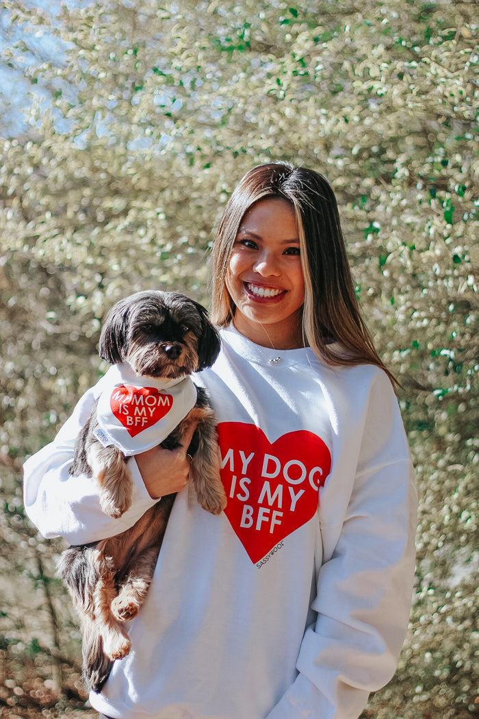 A smiling woman and her dog stand outdoors, both wearing Sassy Woof gear: she wears the unisex “My Dog is my BFF (RED)” sweatshirt, while her dog sports a matching bandana that reads “My mom is my BFF.”.
