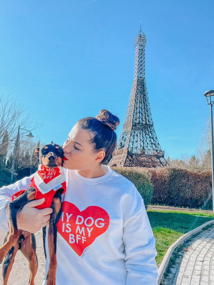 A woman in a white Sassy Woof "Sweatshirt - My Dog is my BFF (RED)" kisses her small dog in a matching outfit, with the Eiffel Tower and clear blue sky behind them. Both feature unisex sizing for the perfect fit.