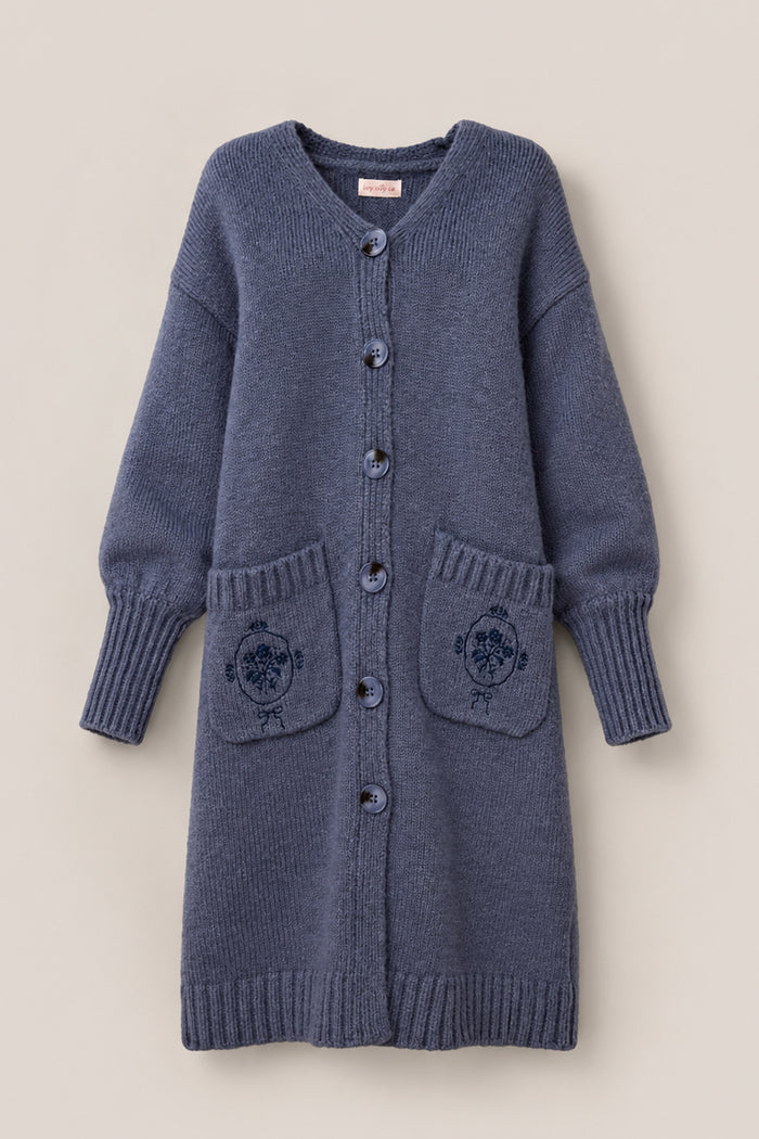 The Teresa Cardigan in Dusty Blue by Ivy City Co is a long, knit sweater featuring large buttons, ribbed cuffs and hem, bishop sleeves, and two front pockets adorned with embroidered circular floral designs.