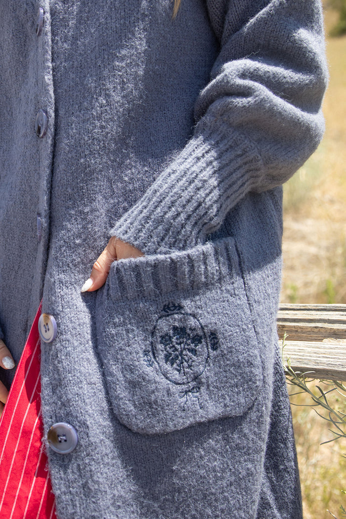 A person wearing the Ivy City Co Teresa Cardigan in Dusty Blue, featuring large buttons and a front pocket with embroidery, stands outdoors near a wooden fence. The cardigan’s subtle bishop sleeves add a touch of elegance as their hand rests on the pocket; red and white striped fabric is visible underneath.