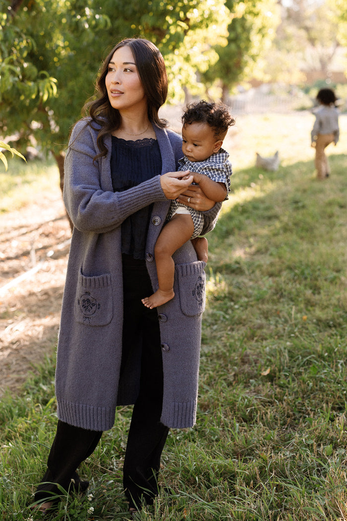 A woman wearing the Ivy City Co Teresa Cardigan in Dusty Blue, featuring elegant bishop sleeves, holds a baby outdoors on a grassy lawn surrounded by trees. Another child walks in the background. The sweater knit cardigan basks in the warm sunlight, enhancing the tranquil scene.