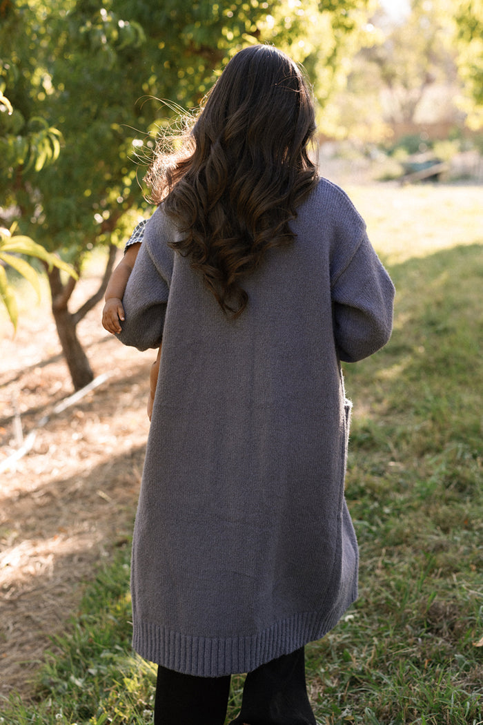 A woman with long wavy hair, wearing the Ivy City Co Teresa Cardigan in Dusty Blue with elegant bishop sleeves, stands outdoors on a grassy path. She is holding a small child on her left side, partially visible, with trees and sunlight in the background.