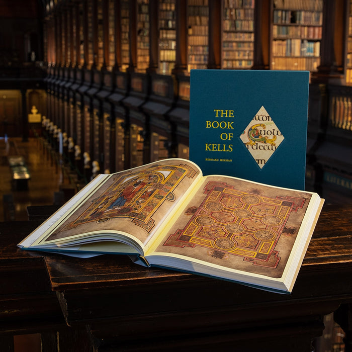 An open book displaying intricate, colorful illustrations from The Book of Kells, placed on a wooden table with bookshelves in the background.