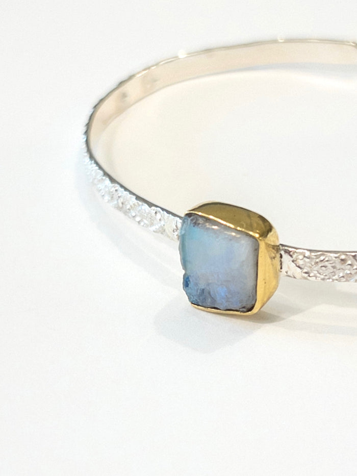 A silver cuff bracelet featuring a rough cut Moonstone gemstone. Moonstone gemstone is set in gold bezel on textured sterling silver cuff