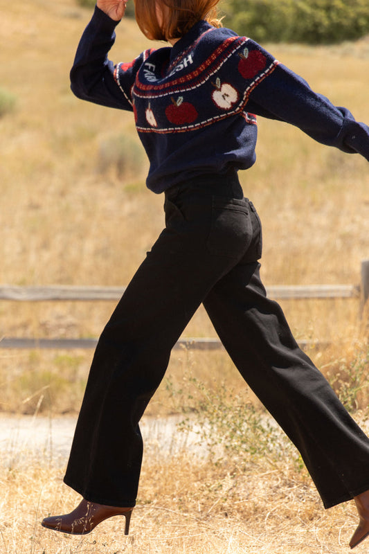 Wearing Ivy City Co’s Tyler Pants in Black and a navy sweater with red and white designs, a woman steps forward outdoors on a dry, grassy path, her arm raised and her head partially out of frame.