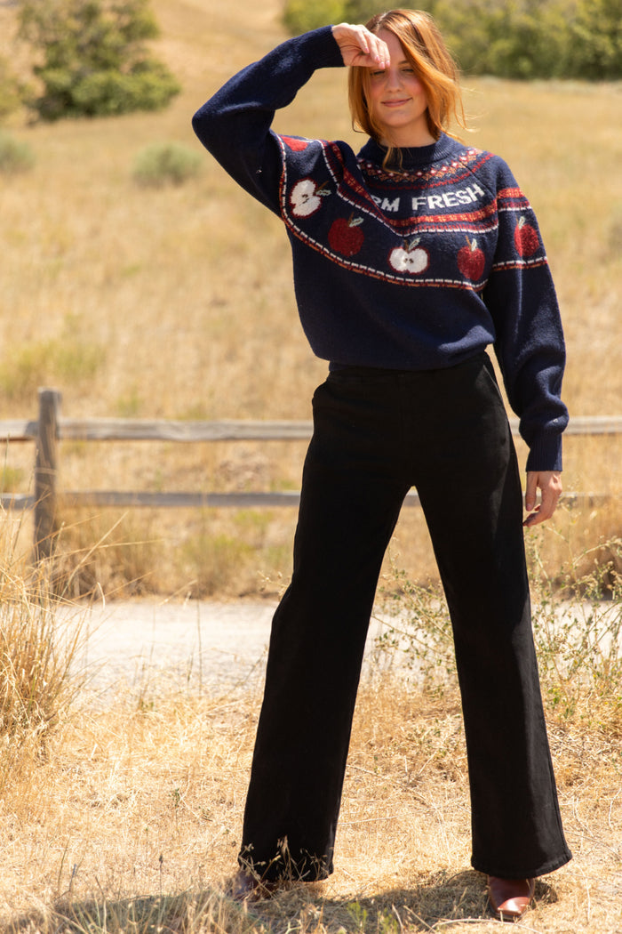 A woman stands outdoors in tall grass, shielding her eyes with one hand. She is wearing Ivy City Co's Tyler Pants in Black and a navy sweater featuring red apple designs and the words FARM FRESH. A wooden fence and dry grass are visible in the background.