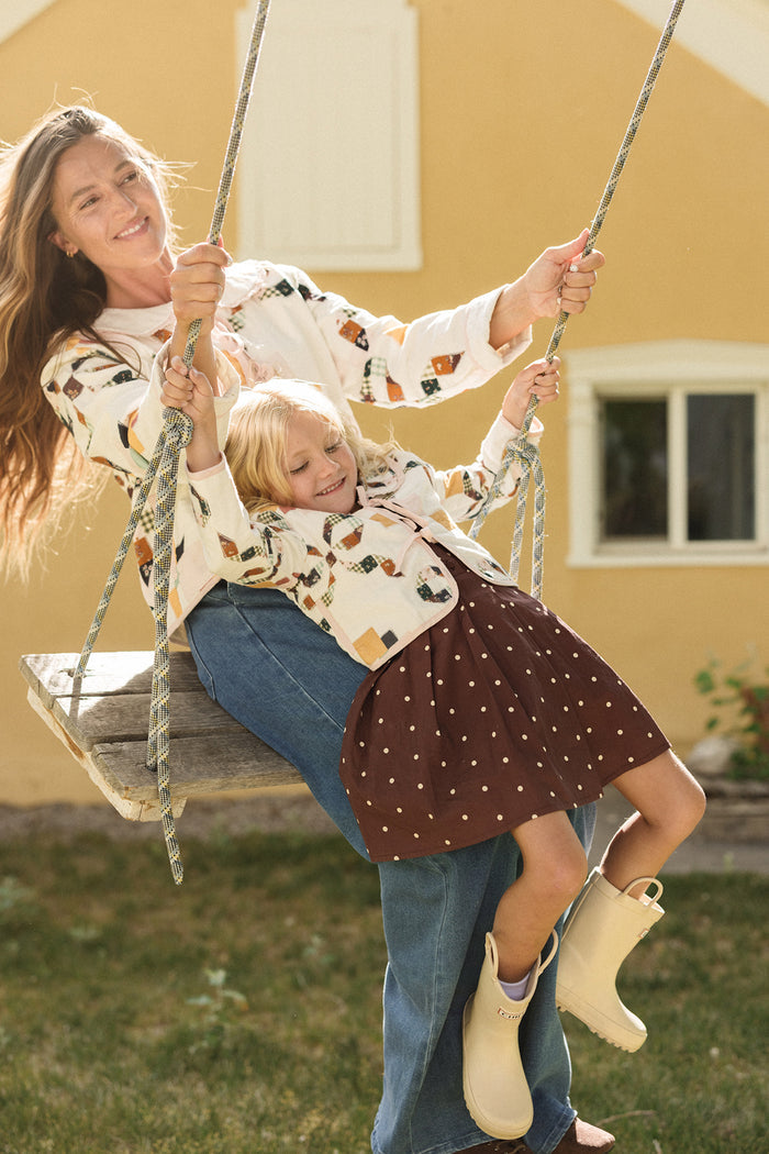 A woman and a young child smile while sitting together on a wooden swing outside a yellow house, enjoying the crisp fall air. The woman, dressed in Ivy City Co’s Tyler Pants in Dark Wash Denim and a patterned jacket, sits beside the child, who wears a brown skirt, light boots, and another cozy patterned jacket.