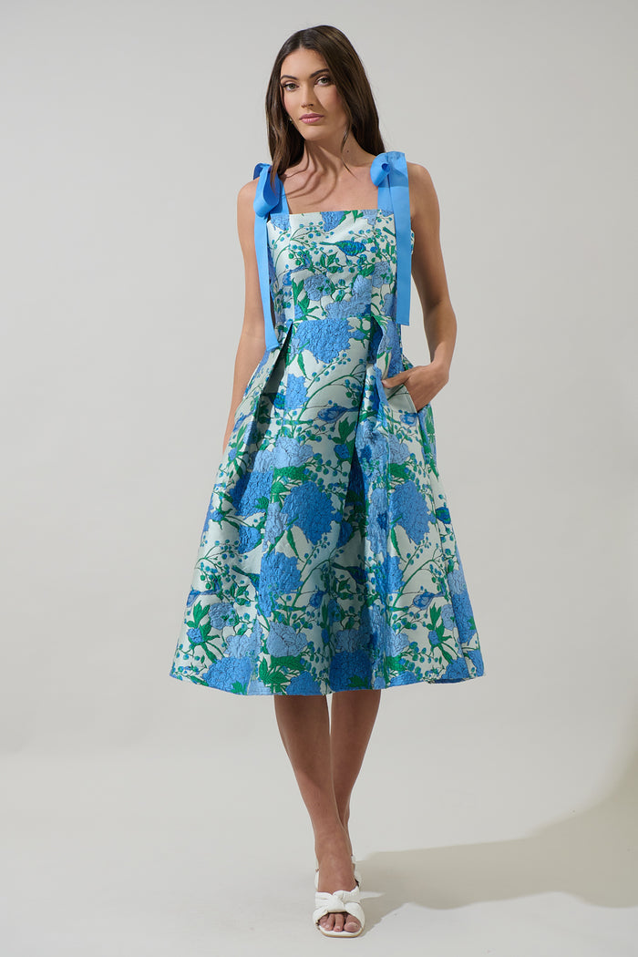 Woman wearing a blue and green floral dress with blue ribbon straps with a plain background