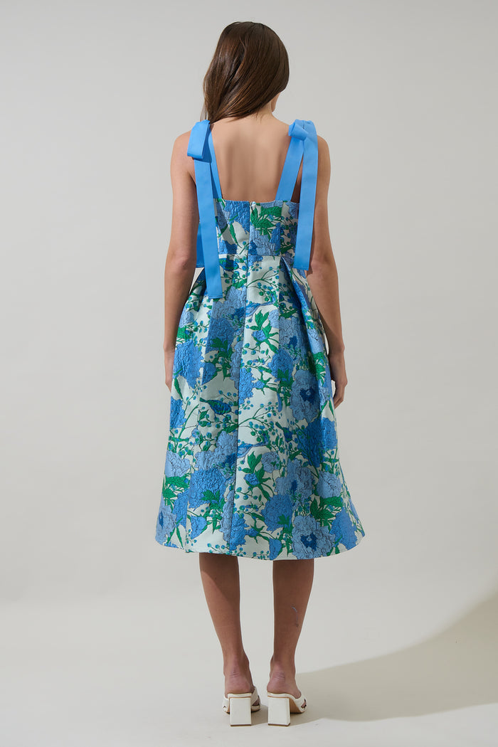 Woman wearing a blue and green floral dress with blue ribbon straps with a plain background
