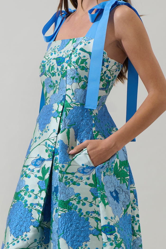 Woman wearing a blue and green floral dress with blue ribbon straps and pockets with a plain background