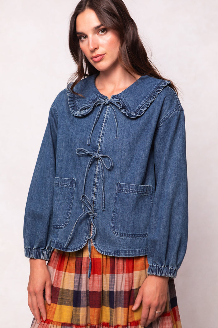 A woman models the Ivy City Co Wynn Jacket in Denim—a maternity and nursing-friendly piece featuring large front pockets, a ruffled collar, and tied bows—styled over a colorful plaid skirt against a light background.