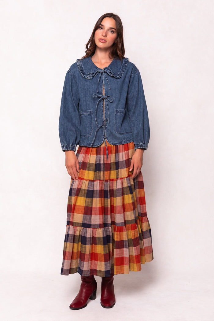 A woman stands against a plain light background wearing the Ivy City Co Wynn Jacket in Denim, featuring a ruffled collar and a maternity nursing-friendly design for both style and comfort. She pairs it with a long multicolored plaid skirt, while her red ankle boots add a pop of color to the ensemble.